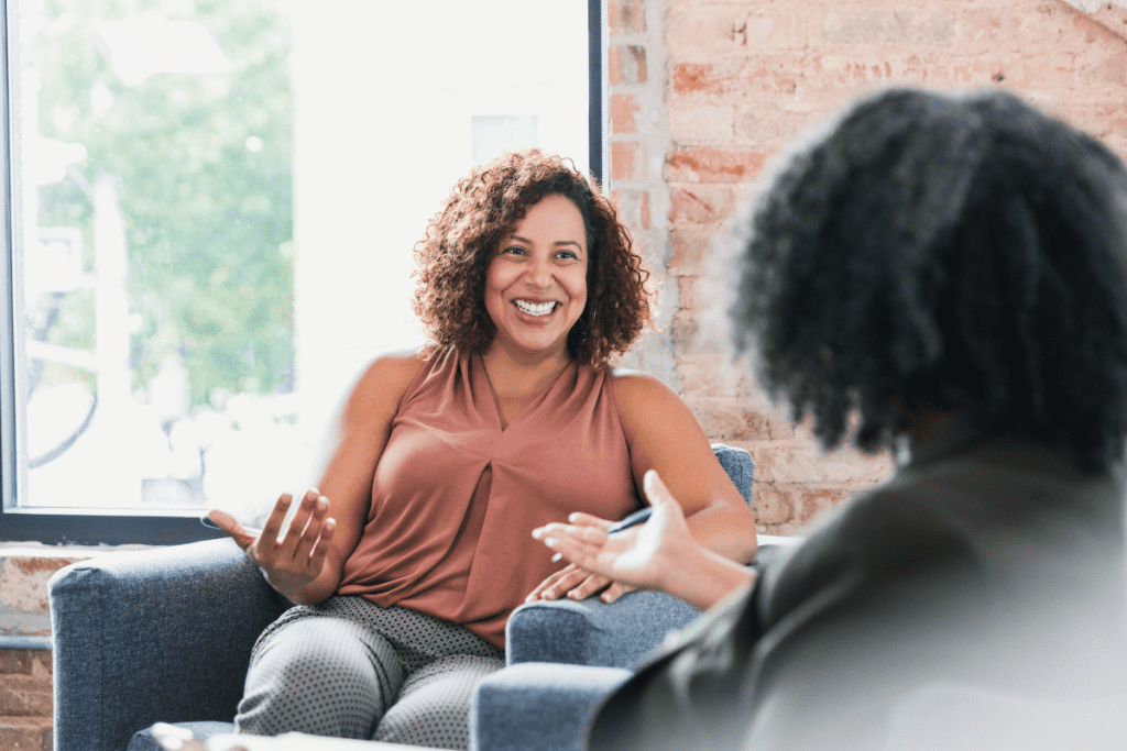 therapist in black shirt talks to client, a female brown woman with curly hair who is smiling while gesturing with her hands. 