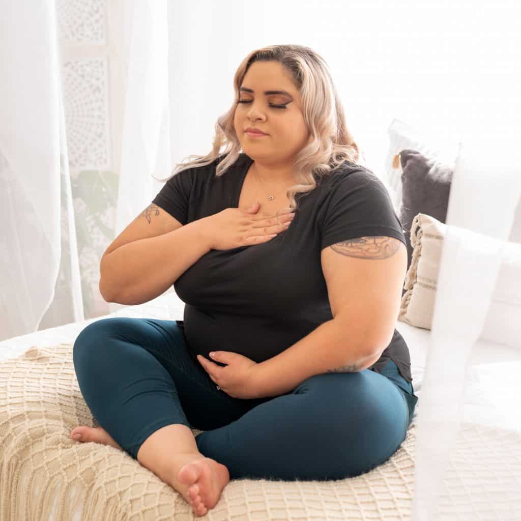 plus size woman sits on bed with leggings meditating with hand on her chest and eyes closed