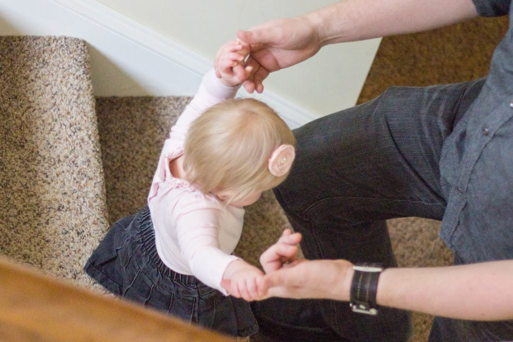 small blonde baby holds her dads hands and walks down stairs