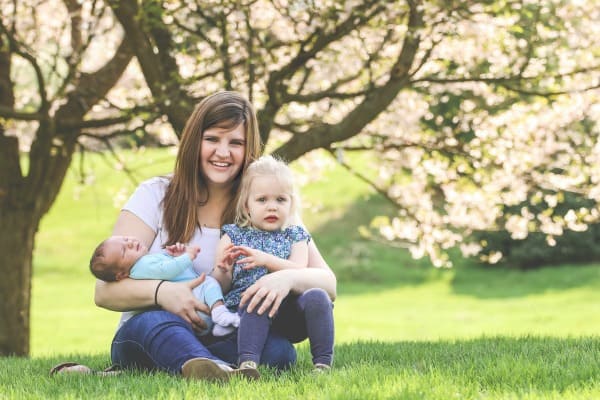 young mom holds her toddler daughter and newborn son under blooming cherry tree
