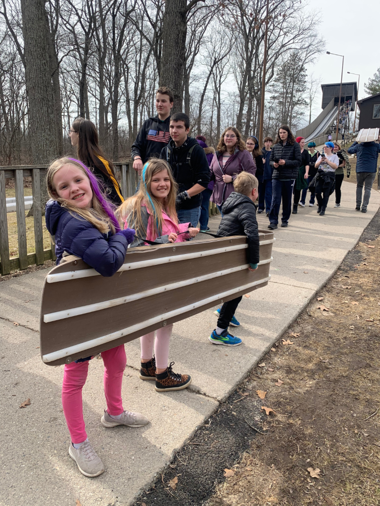 two girls smile big for camera while carrying a toboggan up to a refrigerated slide. A boy helps them but doesn't look at the camera. A crowd of teenagers walk behind them.  