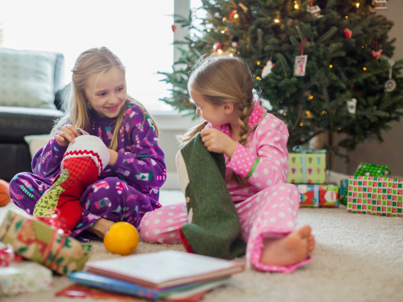 Two elementary age girls open christmas stockings in front of the tree in pink and purple jammies. They are looking at each other with joy and excitement