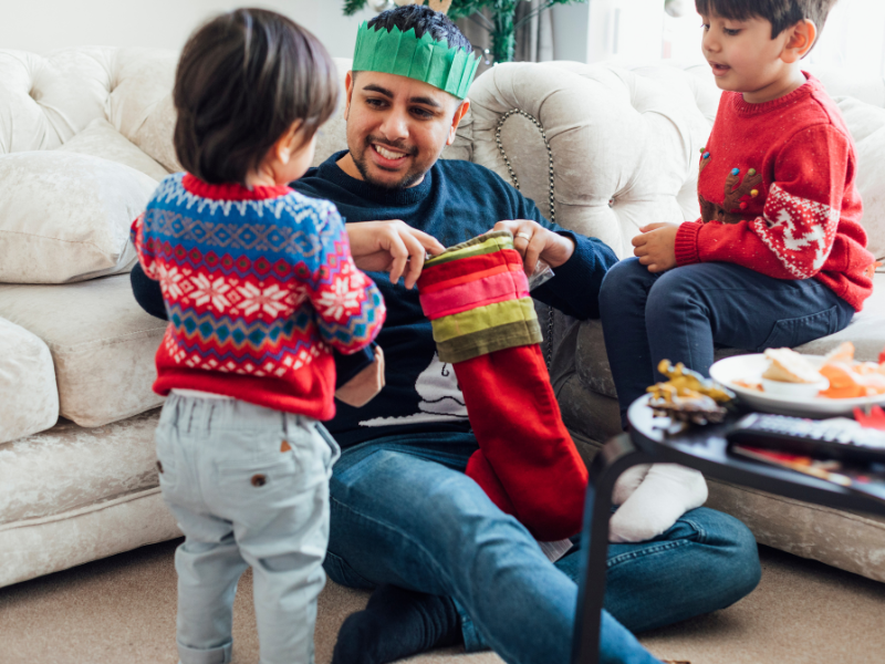 A dad with brown hair and a goatee opens his stocking while his two boy children in christmas sweaters look on. 