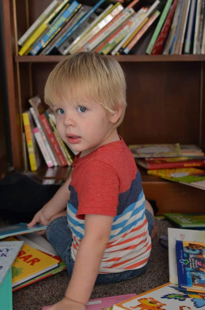 little blonde boy in stripe shirt reads books at overflowing book shelf