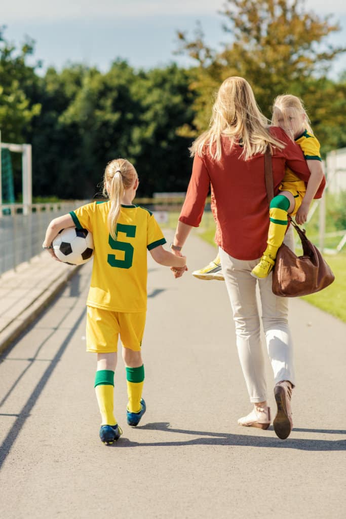 mom in red shirt walks with small blonde soccer player with another kid on her hip