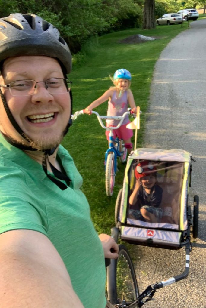 Dad with pull behind bike trailer and son and daughter on small bike take a summer bike ride together