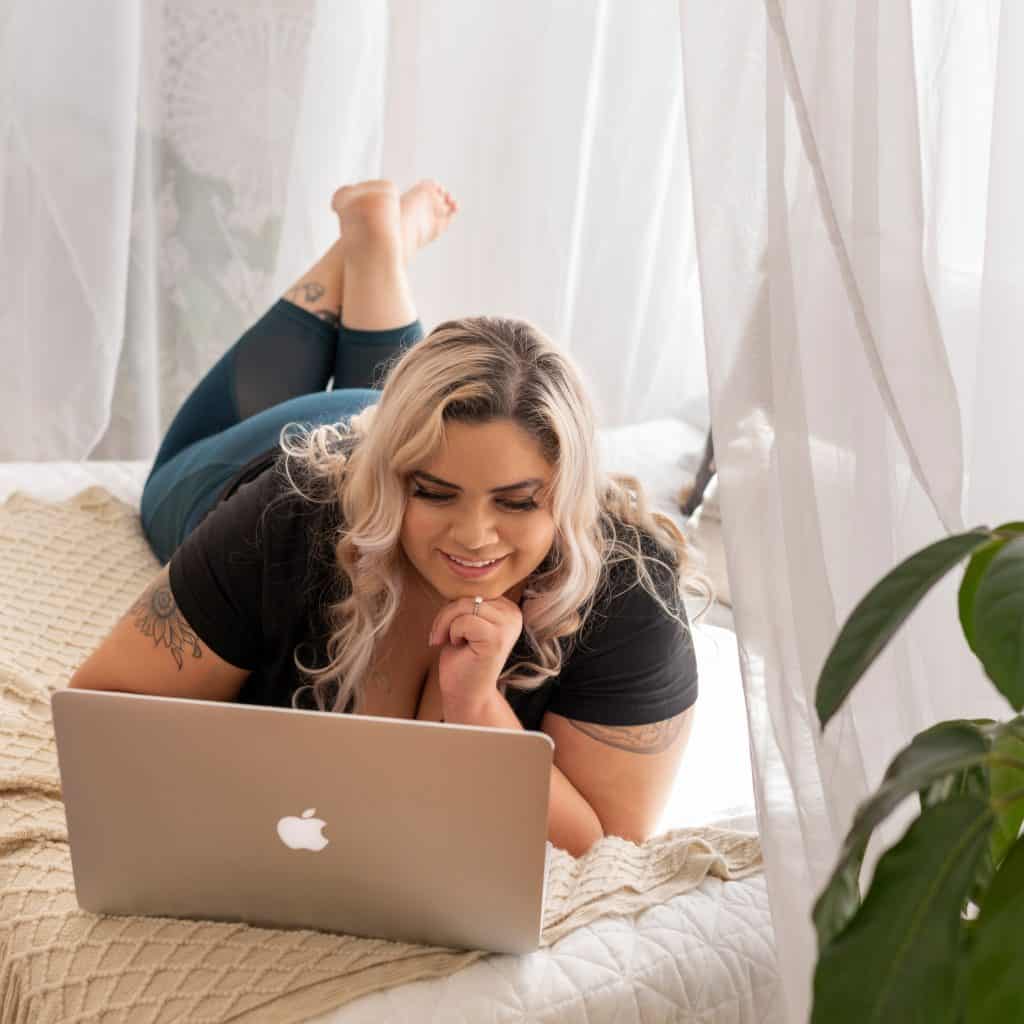 blonde woman with black t-shirt lies on bed looking at laptop