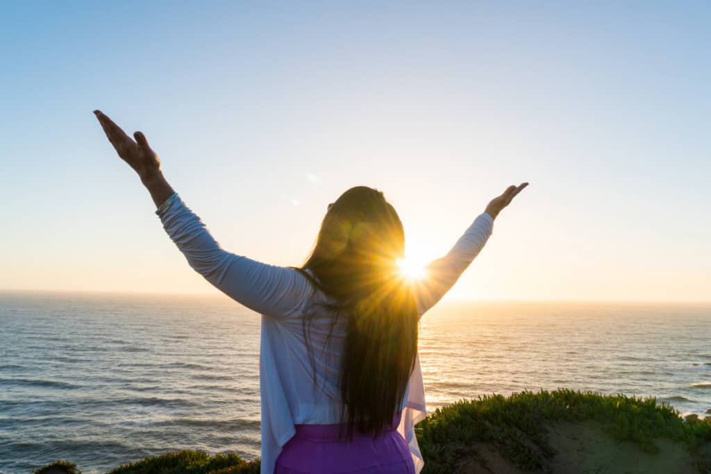 woman with long dark hair raises her hands singing thanks to god while looking at the ocean
