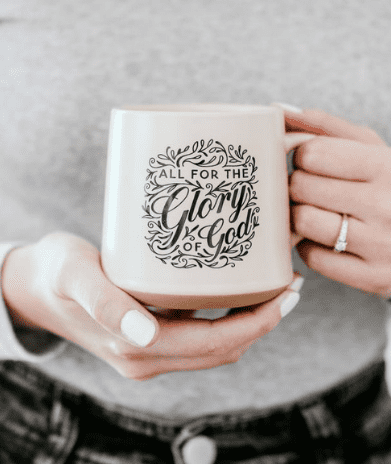 white mug with schroll text all for the glory of God held by woman with white nails in a gray sweatshirt