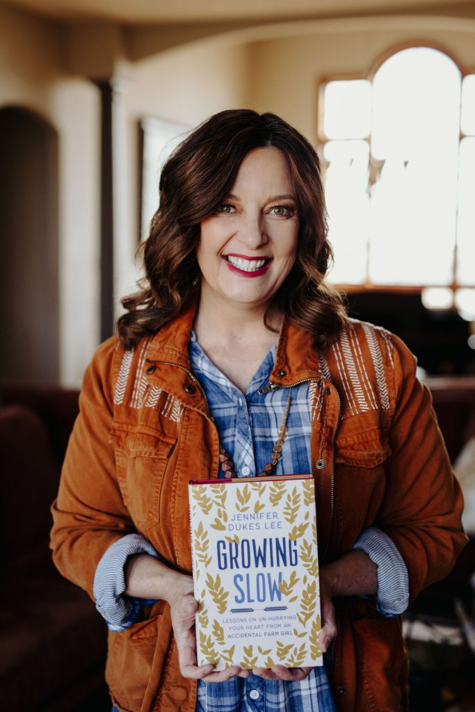 jennifer dukes lee holds her women's Bible study Growing Slow while wearing a leather jacket and plaid shirt in a living room. She is smiling big and backlit by the sunshine.  