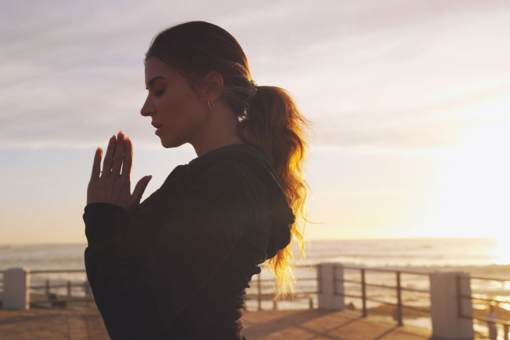 young woman with hair in ponytail pauses to pray as sun sets on a pier