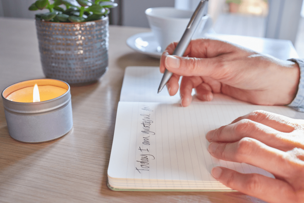 woman holds silver pen over journal writing "things I am grateful for" I plant and small candle are on the table next to her. 
