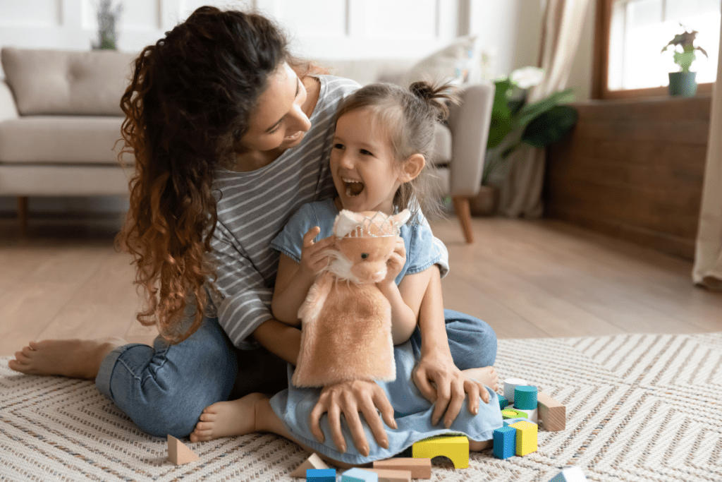 woman with curly brown hair holds small laughing daughter as they play with toys on the floor