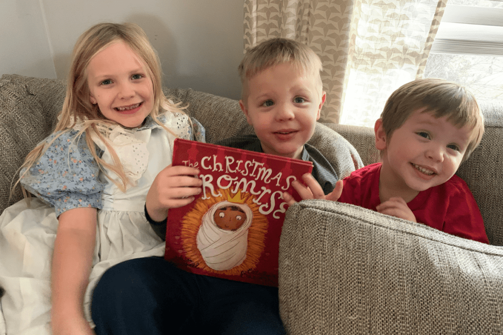 three small children sit on big couch holding the christmas promise book with smiles on their faces