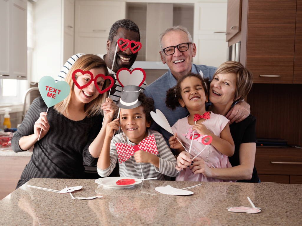 multi-generational and mixed race family gather in a kitchen table holding up home made Valentine's Day cards. You can see the love and joy on their faces. 