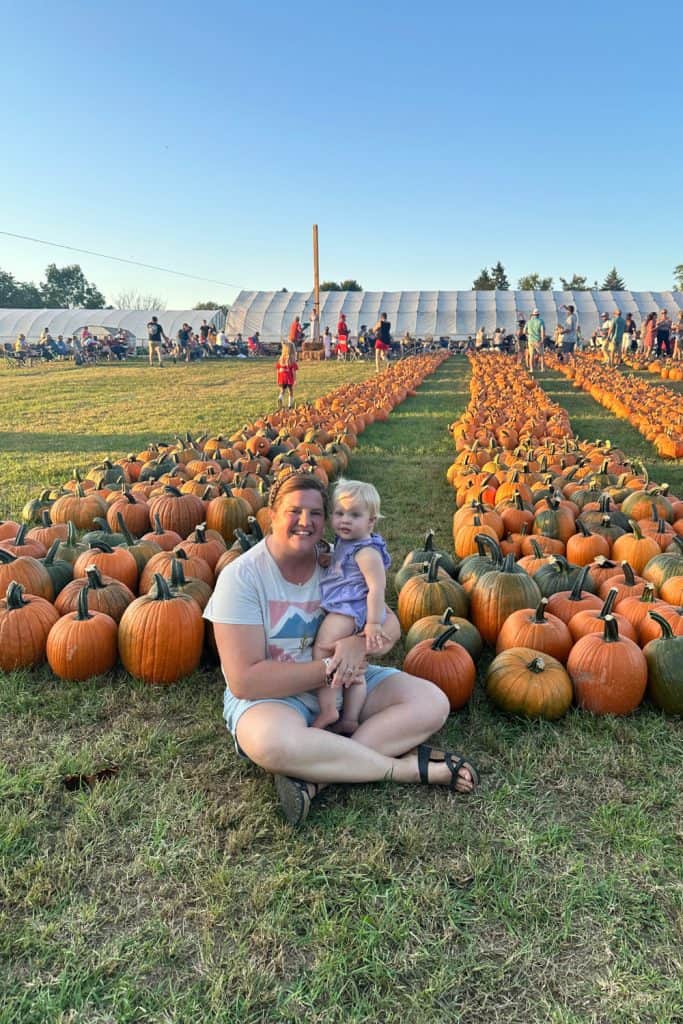 mom sits in front of rows of bright orange pumpkins holding her baby girl on a beautiful fall bucket list day