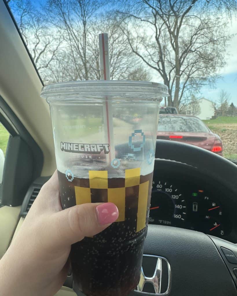 woman with pink nail polish holds a diet coke from Mcdonalds aloft in her mini van in the school pickup line